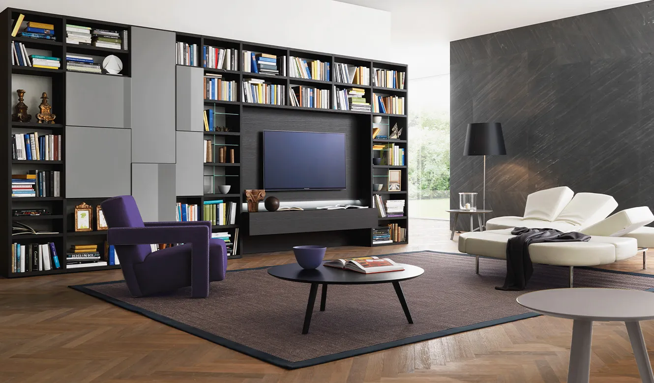 Soggiorno con libreria scura a parete, TV, poltrona viola, chaise longue bianca e tavolino nero su parquet a spina