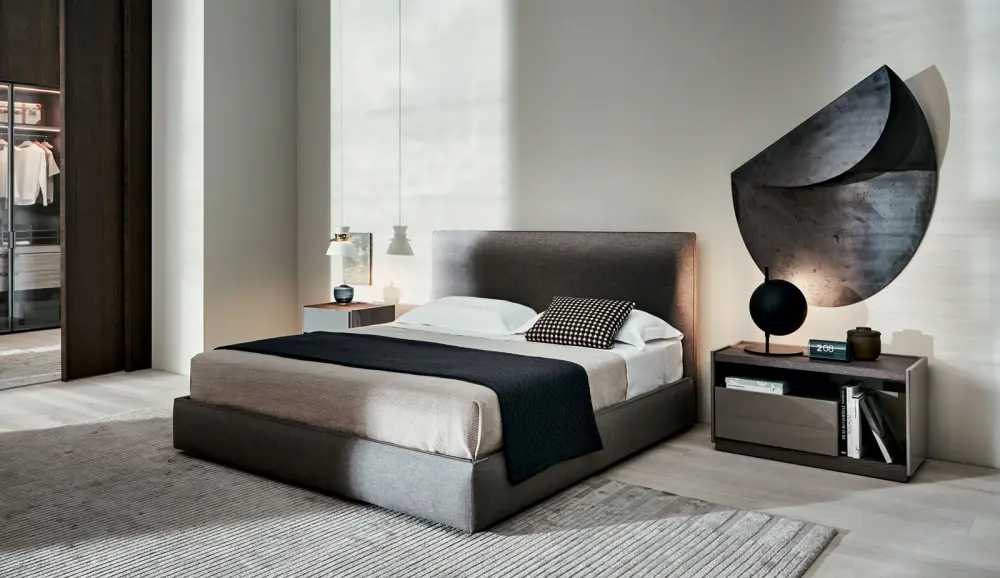 Camera con letto imbottito in tessuto grigio, plaid nero, comodino in legno scuro e scultura metallica a parete